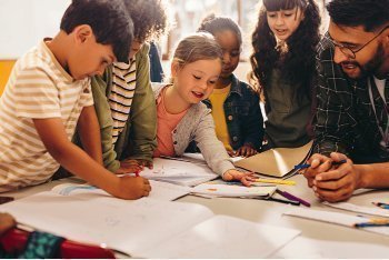 Un docente con gafas guía a un grupo diverso de niños que trabajan con papeles y cuadernos alrededor de una mesa, en un espacio luminoso de aprendizaje colaborativo.