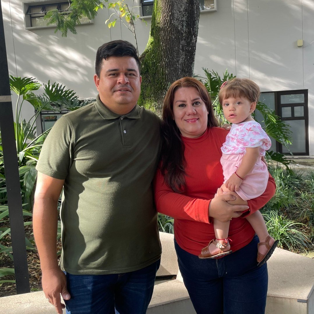 Familia de tres: un hombre a la izquierda con polo verde, una mujer en el centro con su bebé a la derecha; todos sonrientes, en un entorno exterior con plantas y un edificio blanco al fondo.