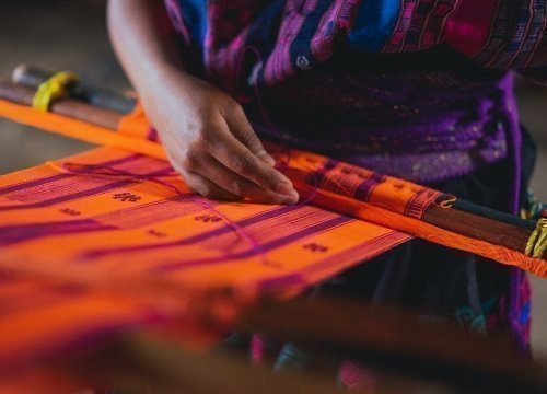 Persona tejiendo a mano un colorido textil tradicional en un telar de cintura, con hilos anaranjados y morados.