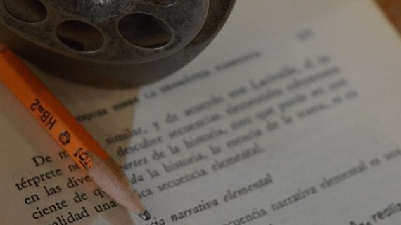 Lápiz naranja sobre un documento escrito, con un objeto circular negro en la esquina superior.