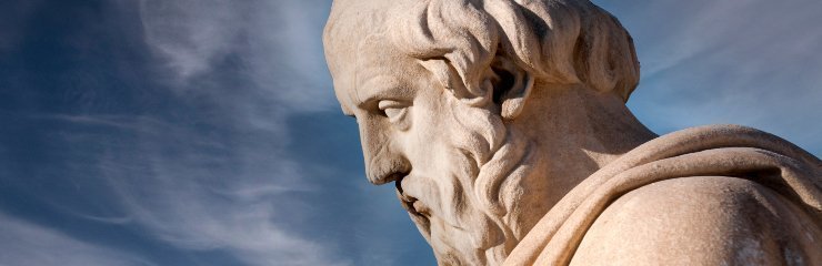 Primer plano de una escultura de un filósofo con barba y expresión pensativa, tallada en piedra clara. El fondo muestra un cielo azul con algunas nubes suaves.