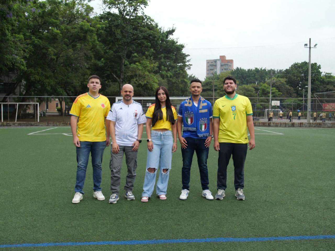 En la imagen se observa a cinco personas de pie sobre una cancha de fútbol sintética, posando frente a la cámara. Cada una viste una camiseta diferente relacionada con selecciones de fútbol internacionales.