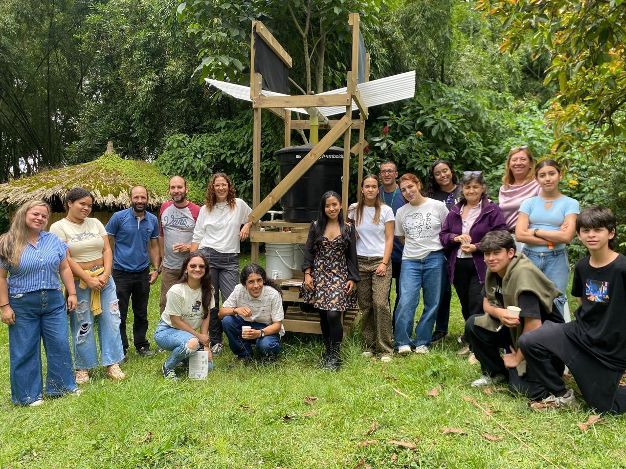 La imagen muestra a un grupo de estudiantes, docentes y miembros de la comunidad reunidos alrededor de una estructura sostenible de almacenamiento y filtración de agua en un entorno natural. El encuentro refleja un trabajo colaborativo enfocado en solucio